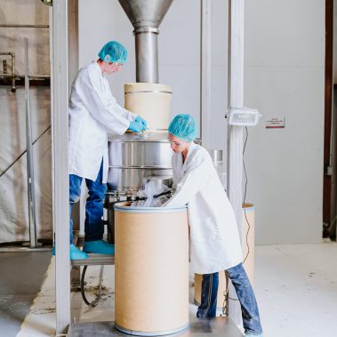 Workers in protective gear operate industrial drying equipment at http://pulsedry.com/, handling large drums during a process involving spray drying systems and powder dryers.