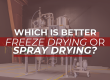 Industrial spray drying equipment inside a manufacturing facility with large metal vessels and pipes. The foreground features a bold headline overlay in white and red text: “Which is better: Freeze Drying or Spray Drying?”—visually highlighting a key processing equipment comparison topic.