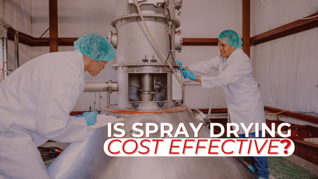 Supporting Blog Is spray drying cost effective Two scientists in lab coats and hairnets working on a large industrial spray dryer. Overlaid white and red text asks: 'IS SPRAY DRYING COST EFFECTIVE?'